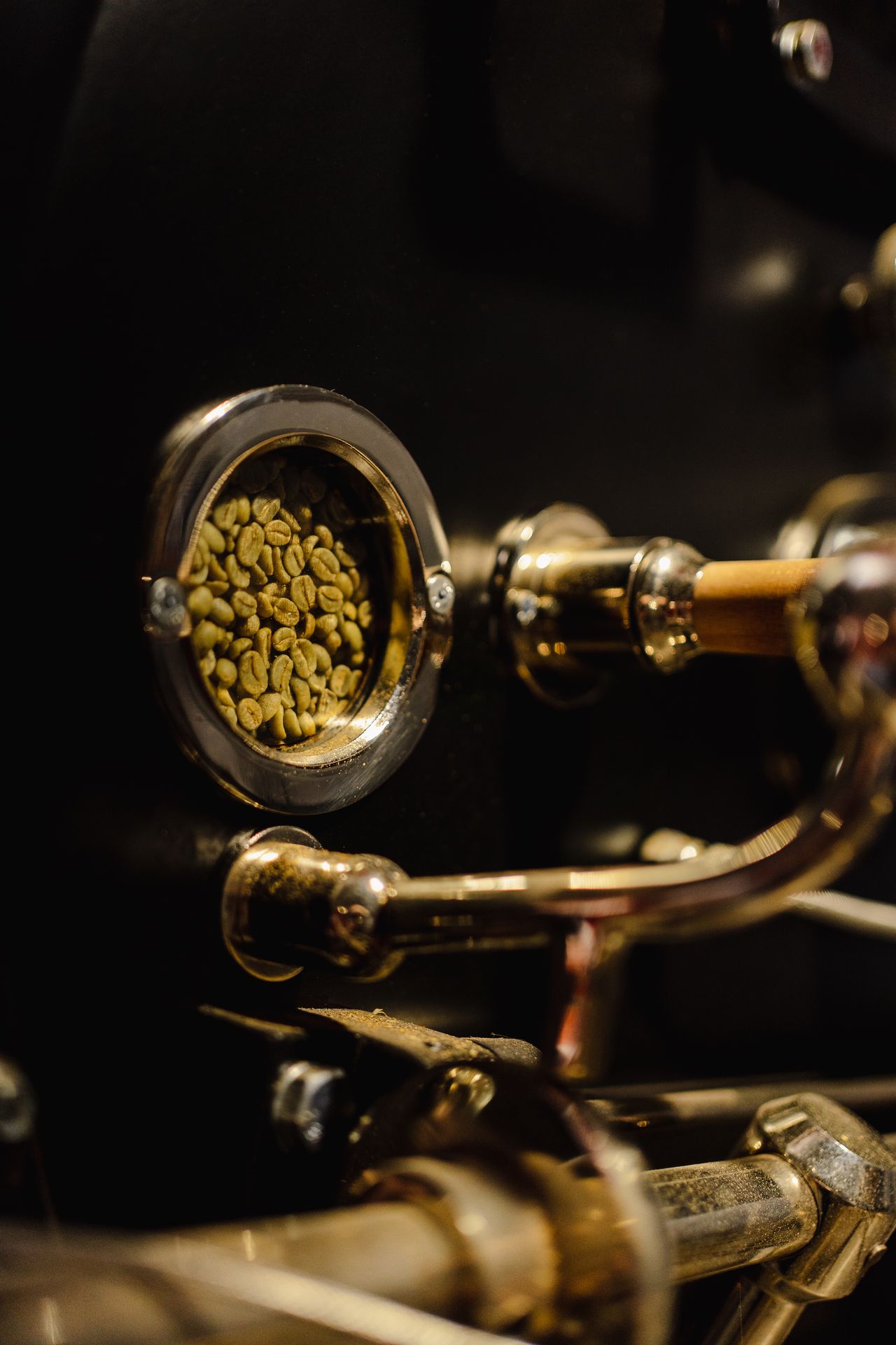 Gros plan sur le tambour d’un torréfacteur à café contenant des grains de café verts, visibles à travers le hublot circulaire en verre. Les grains, encore non torréfiés, présentent leur couleur vert pâle caractéristique, juste avant le début du processus de torréfaction. Les éléments métalliques du torréfacteur, patinés par l’usage, évoquent un savoir-faire artisanal, la précision du geste du torréfacteur et le travail en atelier. Cette image illustre une étape clé de la torréfaction du café de spécialité, reliant le grain vert à la transformation qui révélera les arômes du café.