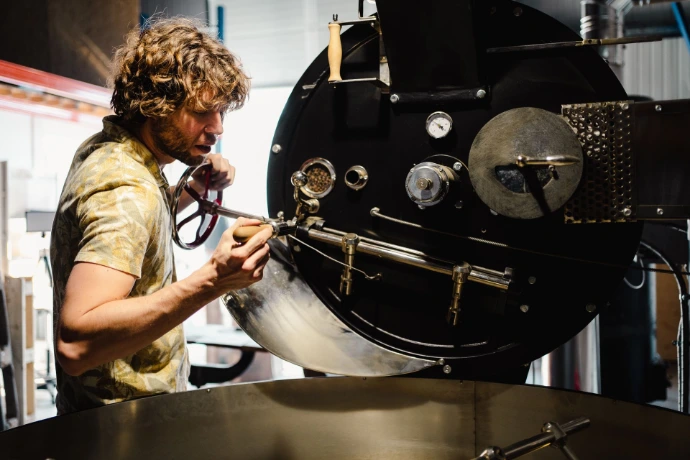 Un torréfacteur artisanal ajuste et contrôle un torréfacteur à café professionnel dans un atelier de torréfaction. L’homme actionne les leviers et instruments de contrôle du tambour, surveillant avec précision le processus de torréfaction des grains de café. Les éléments mécaniques, le métal poli et l’environnement industriel soulignent le savoir-faire du torréfacteur, la maîtrise des profils de torréfaction et l’exigence de qualité propre au café de spécialité. Cette image illustre le travail humain derrière la transformation du café, du grain vert au café torréfié prêt à être dégusté.
