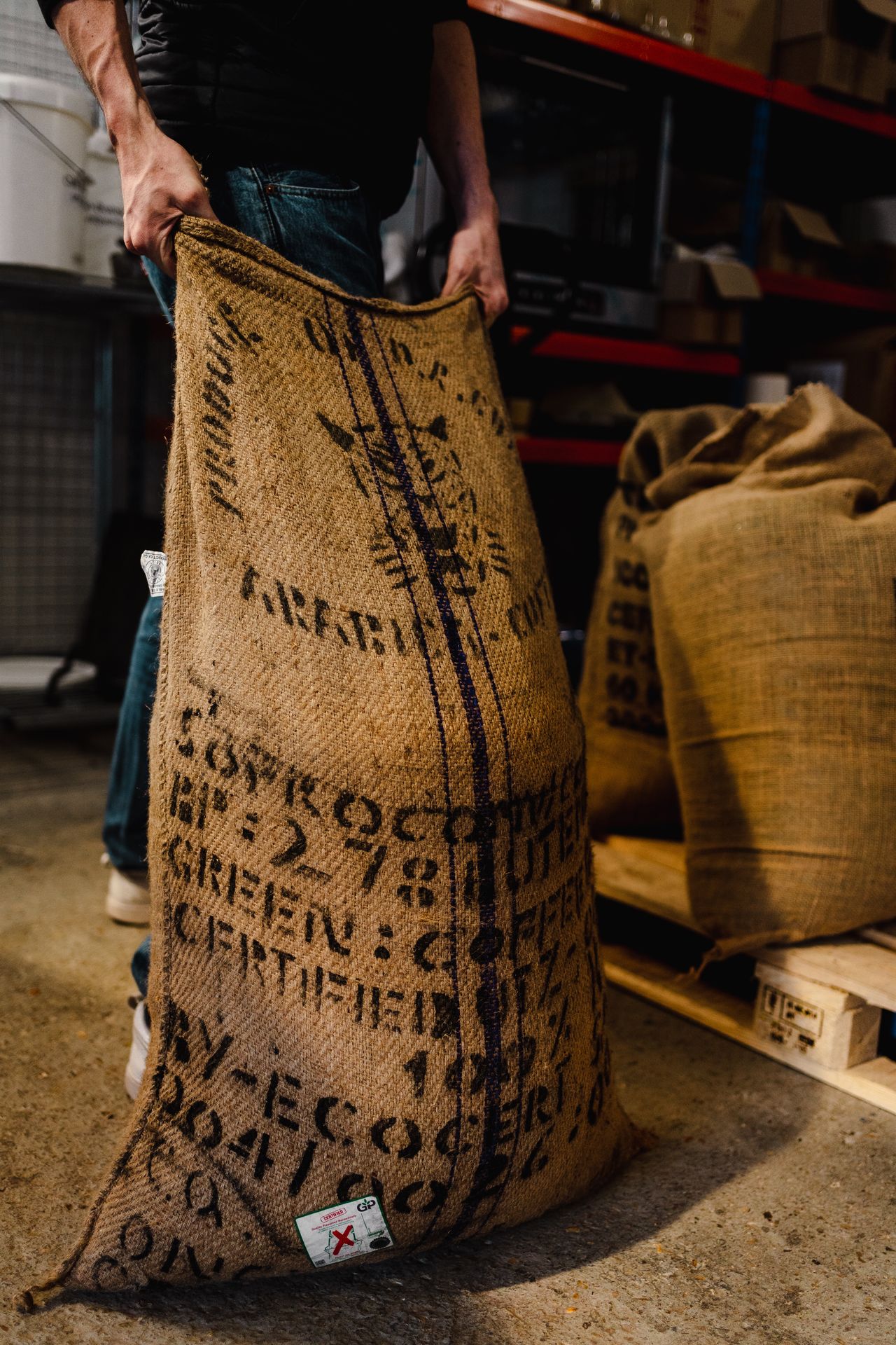 Un sac de café en toile de jute rempli de grains de café vert est manipulé dans un atelier de torréfaction. Le sac, marqué d’inscriptions d’origine et de traçabilité, repose sur un sol industriel, avec d’autres sacs de café empilés à l’arrière-plan. Cette scène illustre l’étape de réception et de stockage du café vert, essentielle dans la filière du café de spécialité. L’image met en avant le travail du torréfacteur, l’importation du café, la traçabilité des lots et l’aspect brut et authentique du métier de torréfacteur artisanal.