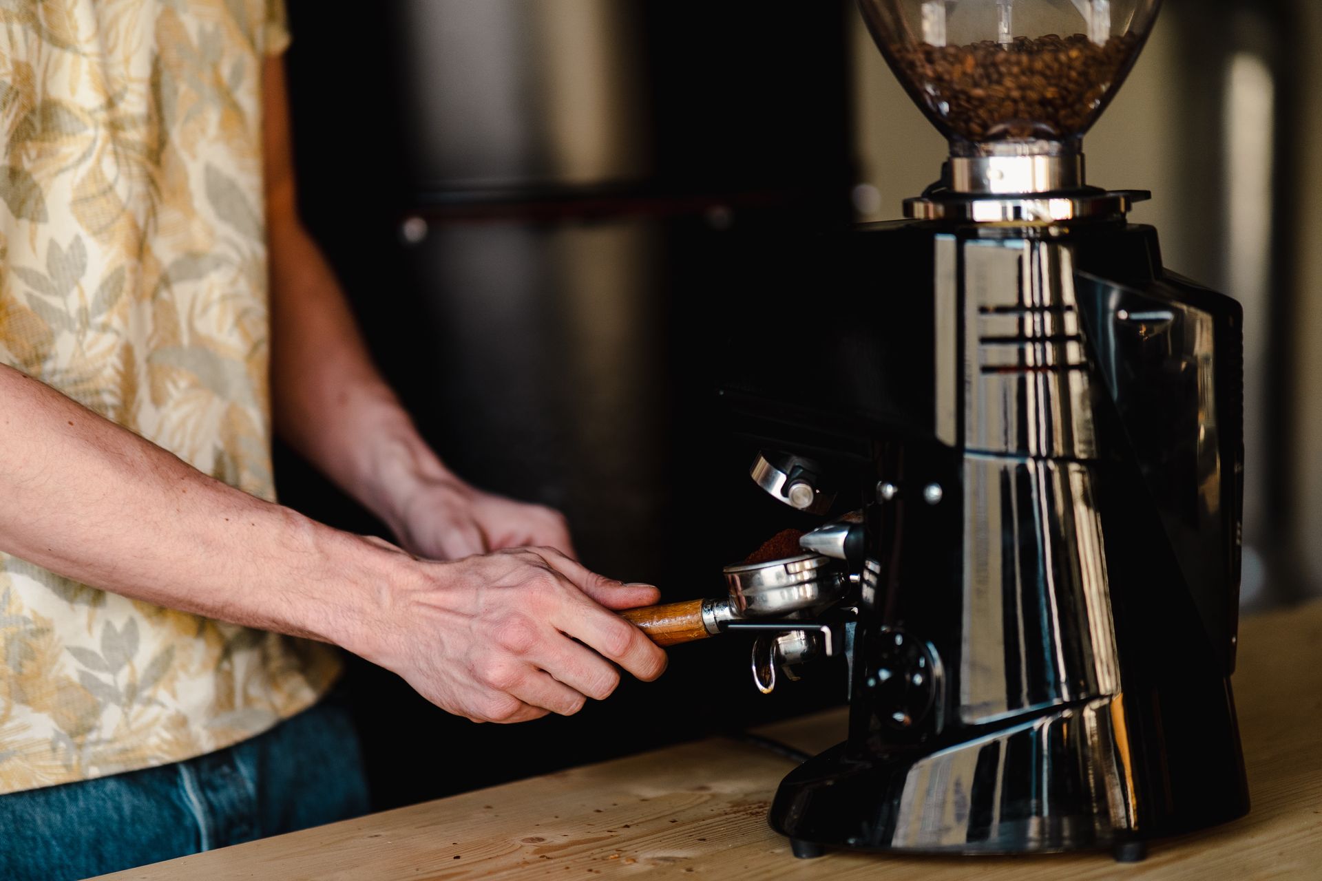 Un barista insère un porte-filtre rempli de café fraîchement moulu dans un moulin à café professionnel, posé sur un plan de travail en bois. Les grains de café torréfiés sont visibles dans la trémie transparente du moulin, soulignant la fraîcheur de la mouture réalisée à la demande. Le geste précis met en avant la préparation du café espresso, le réglage de la mouture et l’importance du matériel dans l’extraction. Cette image illustre le savoir-faire du barista, la culture du café de spécialité et l’attention portée à chaque étape, du grain à la tasse.