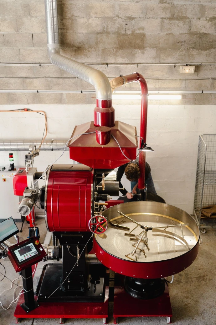 Vue d’ensemble d’un torréfacteur à café industriel de couleur rouge, installé dans un atelier de torréfaction professionnelle. La machine comprend le tambour de torréfaction, un refroidisseur à café circulaire avec bras mélangeurs, un système d’aspiration et de filtration des fumées, ainsi qu’un pupitre de contrôle numérique permettant de piloter précisément les profils de torréfaction. Un torréfacteur intervient sur la machine, illustrant le travail artisanal et technique nécessaire à la transformation du café vert en café torréfié. Cette image met en valeur le savoir-faire, la maîtrise des températures et le rôle central de la torréfaction dans la qualité du café de spécialité.