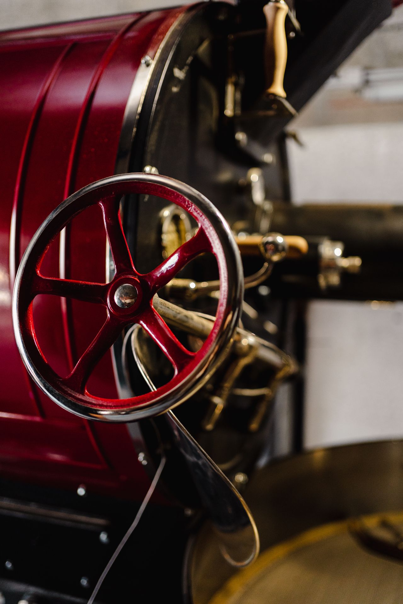 Gros plan sur la manivelle et les éléments mécaniques d’un torréfacteur à café professionnel, peints en rouge profond, symboles du savoir-faire artisanal et de la précision du métier de torréfacteur. Le volant métallique permet le contrôle manuel du tambour et l’accès aux différentes phases de la torréfaction du café. Les détails en métal poli et les poignées en bois soulignent la qualité de fabrication et l’esthétique industrielle de l’équipement. Cette image met en avant la maîtrise du processus de torréfaction, l’attention portée aux réglages et l’importance du geste humain dans la production de café de spécialité.